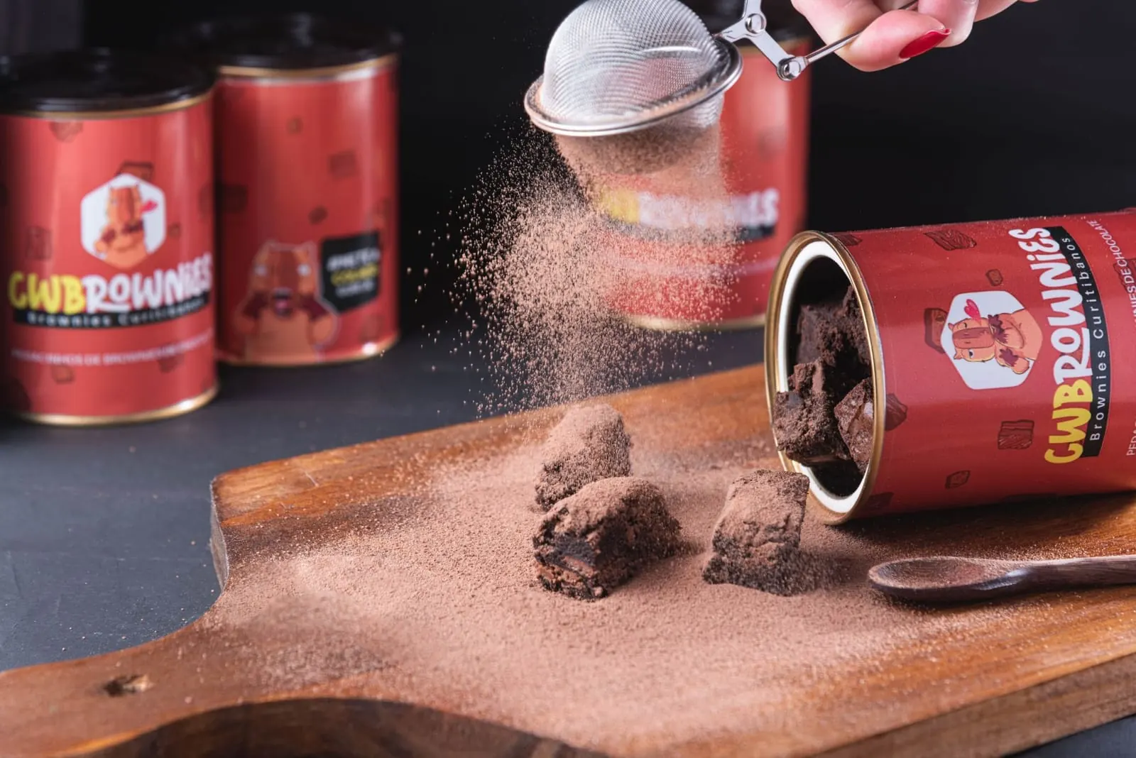 Cacau peneirado sobre pedaços de brownie saindo da lata CWBrownies em tábua de madeira