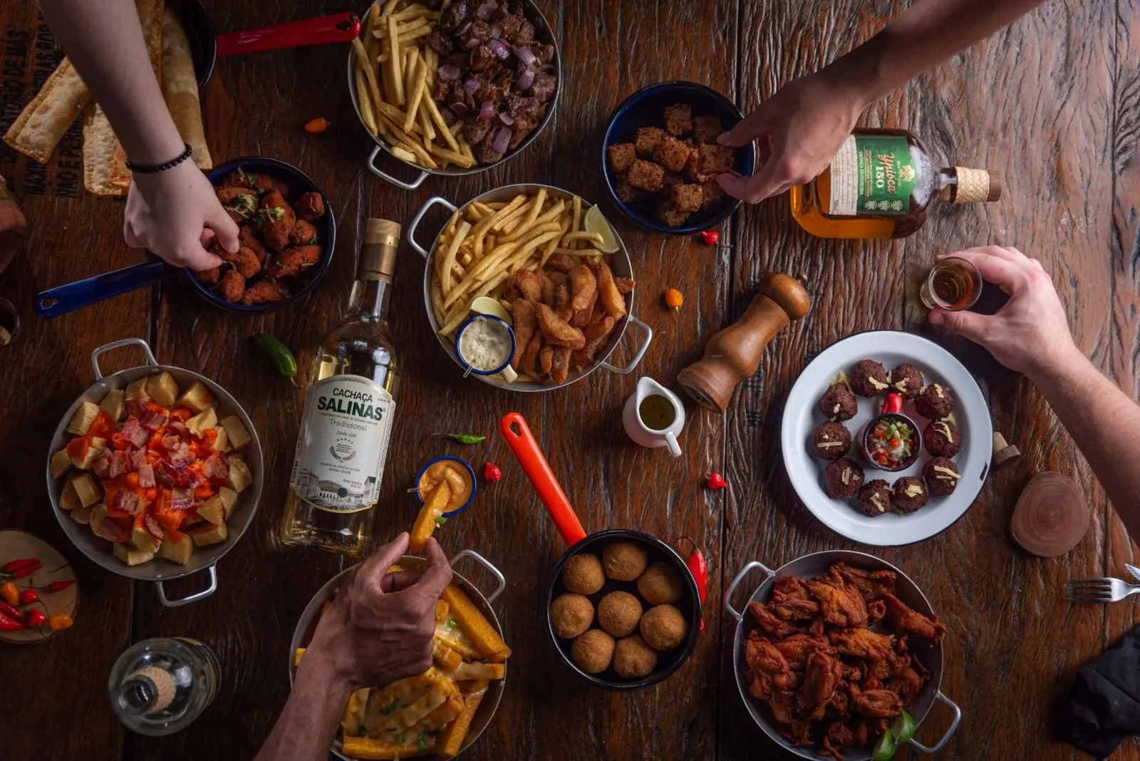 Mesa de boteco vista de cima com porções de petiscos, cachaças e mãos se servindo