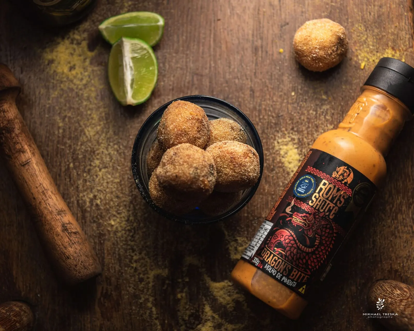Bolinhos empanados em copo com limão e garrafa de molho de pimenta Rom’s Sauce sobre madeira